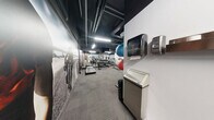 Located at 763 Woodbine Ave in Toronto's East End, this dedicated exercise room delivers a well-equipped fitness experience within a compact, efficiently organized single-level layout. The space features a dark, industrial-style ceiling with exposed ductwork and bright fluorescent strip lighting that creates a focused, energetic atmosphere. Carpeted flooring in a neutral grey tile pattern runs throughout, providing a durable and practical surface underfoot.

The facility is furnished with a solid selection of equipment, including a treadmill, elliptical trainer, stationary cycle, a cable/weight machine, a dumbbell rack with a range of free weights, and an adjustable weight bench — offering both cardio and strength training options. Wall-mounted stability balls and foam rollers provide additional flexibility and recovery tools, while a large floor fan supports air circulation during workouts.

A standout design feature is the large-format athletic photography mural spanning a full wall, depicting dynamic cycling and fitness imagery that adds visual energy and motivation to the space. Full-length mirrors along one wall enhance the sense of openness and allow users to monitor their form. An accessible, gender-neutral washroom is conveniently located within the space. Clean white lower walls contrast with the dark ceiling, giving the room a modern, purposeful aesthetic suited for focused training.