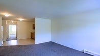 This single-floor residence presents a clean and straightforward layout designed for comfortable, everyday living. The main living area is finished with wall-to-wall carpeting in a neutral tone, creating a warm and quiet underfoot experience. Smooth, white walls and a flat ceiling contribute to a bright, open ambiance throughout the space. A partial dividing wall subtly separates the living area from the kitchen, providing a sense of definition between the two zones while maintaining an open feel. The kitchen is finished with light-colored tile flooring and features warm-toned wooden cabinetry that offers ample storage. A countertop workspace runs along the kitchen wall, making it a practical area for meal preparation. Recessed overhead lighting in the kitchen ensures the area remains well-lit. The overall color palette throughout the space is light and neutral, lending a clean and move-in-ready aesthetic. The simplicity of the design allows for flexible furnishing and personalization, making it a functional and adaptable home for a variety of lifestyles.
