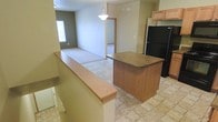 This two-floor space offers a practical and connected layout designed for everyday living. The main level showcases an open-concept design where the kitchen flows naturally into the living area, creating a cohesive and functional environment. The kitchen is equipped with light wood cabinetry, a black refrigerator, and a central island with a warm wood-toned finish that serves as both a workspace and a gathering point. Tile flooring runs throughout the kitchen area, providing a durable and easy-to-maintain surface. A pendant light hangs above, adding a focused point of illumination that complements the overall layout. A half-wall with a wood cap rail defines the transition between the kitchen and the adjacent spaces, lending an architectural detail that adds character without closing off the floor plan. The living area beyond features carpeted flooring and receives natural light through a window, contributing to a comfortable and inviting atmosphere. A doorway leads further into the space, hinting at additional rooms and functionality on this level. The second floor expands the overall footprint of the space, offering additional living or private quarters. Together, the two floors provide a well-rounded layout suited for a variety of uses.