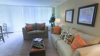 This single-floor space offers a warm and inviting atmosphere, thoughtfully designed for comfort and functionality. The living area serves as the heart of the space, showcasing a soothing color palette of soft greens and warm neutrals that create a calming ambiance throughout. Large windows dressed with vertical blinds allow generous natural light to fill the room, enhancing the open and airy feel. The seating arrangement features a plush sofa and a matching loveseat, both accented with warm orange and patterned throw pillows that add a welcoming pop of color and texture. A tufted ottoman-style coffee table anchors the seating area, providing both a functional surface and a stylish focal point. Subtle decorative touches, including a table lamp, framed wall art, and a lush indoor plant, contribute to the lived-in charm of the space. Carpeted flooring runs throughout the living area, adding softness underfoot and contributing to the overall cozy character of the room. The layout is efficient and well-proportioned, making excellent use of the available space while maintaining an uncluttered, comfortable environment suitable for everyday living and relaxed entertaining.