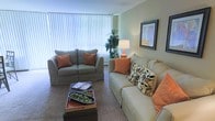 This single-floor space offers a warm and inviting atmosphere, thoughtfully designed for comfort and functionality. The living area serves as the heart of the space, showcasing a soothing color palette of soft greens and warm neutrals that create a calming ambiance throughout. Large windows dressed with vertical blinds allow generous natural light to fill the room, enhancing the open and airy feel. The seating arrangement features a plush sofa and a matching loveseat, both accented with warm orange and patterned throw pillows that add a welcoming pop of color and texture. A tufted ottoman-style coffee table anchors the seating area, providing both a functional surface and a stylish focal point. Subtle decorative touches, including a table lamp, framed wall art, and a lush indoor plant, contribute to the lived-in charm of the space. Carpeted flooring runs throughout the living area, adding softness underfoot and contributing to the overall cozy character of the room. The layout is efficient and well-proportioned, making excellent use of the available space while maintaining an uncluttered, comfortable environment suitable for everyday living and relaxed entertaining.