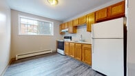 This single-floor unit offers a clean, functional layout with a cohesive design carried throughout by grey-tone laminate wood flooring. The interior is finished with neutral walls and white trim, creating a bright, understated aesthetic that suits a variety of furnishing styles.

The kitchen is well-equipped with warm oak cabinetry extending to the upper and lower walls, providing ample storage space. A white electric range, a stainless steel sink, white refrigerator, and an overhead range hood complete the practical setup. A window above the kitchen area allows natural light to fill the space.

The living and bedroom areas are generously proportioned, each featuring baseboard heating units and windows that bring in natural light and views of the surrounding exterior. The rooms offer flexible, open layouts suitable for varied uses.

A full bathroom includes a soaking tub with a white surround featuring built-in shelving, a toilet, and an oak vanity with a light countertop. Recessed lighting illuminates the space cleanly.

A central hallway connects the rooms efficiently, with a mirrored panel adding depth near the entry. The overall layout, as seen in the floor plan, is compact yet well-organized, maximizing usability across all areas.