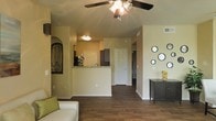 This single-floor residence offers a well-designed open-concept layout that connects the living area and kitchen, creating a spacious and inviting atmosphere. The living space features rich, dark hardwood-style flooring that runs throughout, providing a warm and cohesive aesthetic. A ceiling fan with a light fixture anchors the living area, offering both style and functionality.

The kitchen is defined by a peninsula with granite-style countertops, providing additional prep space and a natural divide between the cooking and living areas. Dark cabinetry in the kitchen adds a contemporary contrast to the warm, neutral-toned walls that carry throughout the space.

A hallway extending from the main living area leads to additional rooms, suggesting a practical and efficient floor plan. The walls are painted in soft, warm tones that enhance the welcoming ambiance. Decorative touches, such as an arched niche with ornamental detail on one wall and a series of circular mirrors on another, add personality and visual interest to the space.

Overall, the residence strikes a balance between functional design and comfortable living, with consistent flooring, cohesive color tones, and thoughtful architectural details that give the space a polished, well-appointed character.