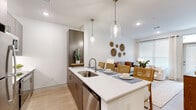 Located at 13959 Hillcroft Ave in Houston, TX, this one-bedroom, one-bathroom residence offers a thoughtfully designed single-floor layout that maximizes every square foot. The open-concept kitchen and living area form the heart of the home, where the kitchen impresses with dark wood cabinetry, stainless steel appliances, and a white quartz peninsula island with seating — illuminated by stylish pendant lighting. Light wood-style flooring runs throughout, lending warmth and continuity to the space. The living room is bright and inviting, featuring large windows with blinds and floor-length curtains that allow natural light to fill the room, complemented by recessed lighting above. The bedroom offers a comfortable and serene retreat with windows providing natural light, a ceiling fan, and ample space for furnishings. A dedicated closet adds practical storage. The bathroom is cleanly appointed with dark wood cabinetry, a white countertop vanity, a large mirror with contemporary lighting fixtures, and light wood flooring. In-unit laundry connections are also visible, adding a layer of everyday convenience. The overall aesthetic is modern and neutral, with crisp white walls creating a clean, airy ambiance throughout.