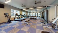 This fitness center is designed to support a comprehensive workout experience within a single, open floor layout. The space is finished with a distinctive blue and tan checkered carpet tile flooring that adds a clean, modern aesthetic throughout. Large windows line multiple walls, flooding the interior with natural light and offering views of the surrounding building exterior, while ceiling fans overhead ensure comfortable air circulation during workouts.

The cardio zone features a row of treadmills, elliptical trainers, and a stationary recumbent bike, all positioned to take advantage of the natural light from the windows. A wall-mounted television is conveniently located nearby for entertainment during cardio sessions. The strength training area is well-appointed with a rack of dumbbells, a weight bench, and multiple cable and multi-press machines, providing options for both isolation and compound exercises. Mirror panels along the walls enhance the sense of space and allow users to monitor their form.

A dedicated functional training corner houses an assortment of stability and medicine balls neatly stored on a rack, supporting flexibility and core conditioning workouts. Pendant and linear fluorescent lighting supplements the natural light, ensuring the space remains well-lit at all hours. The overall ambiance is clean, functional, and inviting, making this fitness center a well-rounded facility for a full-body training routine.