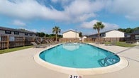 Located in Charleston, SC, Castlewood Townhomes offers a well-maintained community amenity area centered around a generously sized outdoor swimming pool. The pool features a curved, kidney-like shape with clearly marked depth indicators, making it accessible for a range of swimmers. Surrounding the pool is a spacious concrete deck furnished with multiple lounge chairs and chaise lounges, providing ample space for sunbathing and relaxation. Tall palm trees frame the area, lending a resort-like atmosphere that complements the warm South Carolina climate. The pool area is enclosed by a wooden privacy fence, creating a comfortable and secure environment for residents. A classic lamp post adds a charming touch to the setting. The broader community is visible in the background, with multi-unit residential buildings lining the perimeter. The open sky above allows for abundant natural light throughout the day, enhancing the bright and inviting atmosphere of this outdoor space. Overall, the community pool area at Castlewood Townhomes serves as an attractive and functional gathering space for residents seeking leisure and recreation in a pleasant outdoor environment.