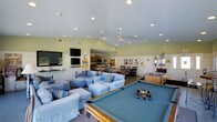 This single-floor community room is a versatile, well-appointed gathering space designed to accommodate a variety of social and recreational activities. The room is anchored by a full-size pool table with a blue felt surface, complemented by a foosball table nearby, making it a hub for game enthusiasts. A comfortable lounge area features plush blue sofas and armchairs arranged around wall-mounted flat-screen televisions, creating an inviting space for relaxed conversation or casual viewing. Vaulted ceilings fitted with recessed lighting and a ceiling fan give the room an open, airy feel throughout. The soft green walls contribute to a bright and cheerful ambiance. Toward the back, a dining area with tables and chairs provides ample seating, seamlessly connected to a kitchenette equipped for light food preparation and serving. Framed artwork and decorative accents on the walls add a personalized touch to the space. The thoughtful layout balances active recreation with comfortable relaxation, making this community room a well-rounded amenity suited for gatherings, events, and everyday use by residents.