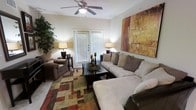 This single-floor residence offers a thoughtfully designed living space that balances comfort and style. The living room serves as the focal point of the home, showcasing a generously sized sectional sofa complemented by a coordinating armchair, both upholstered in rich, earthy tones. A large, bold abstract canvas in warm shades of amber, rust, and gold anchors one wall, adding a striking artistic element to the room. Crown molding frames the ceiling, where a classic five-blade ceiling fan with an integrated light fixture provides both comfort and subtle elegance. Natural light filters in through a set of French doors fitted with blinds, creating a bright and welcoming atmosphere. The flooring is accented by a multi-toned geometric area rug in green, red, and tan hues, adding texture and visual interest to the space. A dark wood coffee table centered in the seating area provides a practical and stylish surface, while coordinating table lamps on either side of the room cast a warm, ambient glow. Lush indoor greenery adds a refreshing natural touch, completing the inviting and well-balanced aesthetic of this comfortable living space.