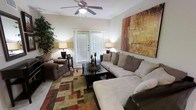 This single-floor residence offers a thoughtfully designed living space that balances comfort and style. The living room serves as the focal point of the home, showcasing a generously sized sectional sofa complemented by a coordinating armchair, both upholstered in rich, earthy tones. A large, bold abstract canvas in warm shades of amber, rust, and gold anchors one wall, adding a striking artistic element to the room. Crown molding frames the ceiling, where a classic five-blade ceiling fan with an integrated light fixture provides both comfort and subtle elegance. Natural light filters in through a set of French doors fitted with blinds, creating a bright and welcoming atmosphere. The flooring is accented by a multi-toned geometric area rug in green, red, and tan hues, adding texture and visual interest to the space. A dark wood coffee table centered in the seating area provides a practical and stylish surface, while coordinating table lamps on either side of the room cast a warm, ambient glow. Lush indoor greenery adds a refreshing natural touch, completing the inviting and well-balanced aesthetic of this comfortable living space.