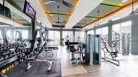 Located at Post Oak Park Drive in the heart of Houston, TX, this professionally designed fitness center delivers a premium workout experience across a generously sized single-floor layout. The space is defined by its striking floor-to-ceiling windows that wrap around the perimeter, flooding the interior with abundant natural light and offering expansive views of the Houston skyline and surrounding urban landscape.

The interior design features a contemporary aesthetic, with a coffered ceiling accented by warm wood tones and vibrant lime-green trim details that add energy to the space. Multiple ceiling fans are suspended throughout, ensuring comfortable airflow during workouts. Light wood-plank flooring runs throughout, complementing the modern gray and green color palette.

The exercise room is comprehensively equipped with a full suite of Matrix-brand machines, including cable systems, strength-training stations, treadmills, stair climbers, stationary bikes, and rowing machines. A dedicated free weights area includes a full dumbbell rack, barbells, benches, and floor mats for functional training. Wall-mounted televisions provide entertainment throughout the space. An adjacent glass-walled studio is visible from the main floor, offering additional programming space. The overall ambiance is bright, motivating, and well-organized, making this fitness center a standout amenity.