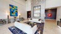 This striking lobby is designed to make a bold architectural statement from the moment of arrival. Floor-to-ceiling glass walls at the entrance flood the space with natural light while offering views of the surrounding exterior. Polished marble-style tile flooring extends throughout, lending an air of refinement and continuity across the entire level.

The central lounge area serves as the focal point, anchored by a pair of sleek white sofas arranged around a glass-top coffee table, all set atop a richly patterned area rug in deep blues and earth tones. A dramatic textured accent wall with a wave-relief design rises to the full height of the space, complemented by an oversized mirror that amplifies the sense of openness. Above, a sculptural chandelier with branching metallic arms adds a distinctive artistic touch.

Vibrant large-scale paintings enliven the walls, contributing to a curated gallery-like atmosphere. A dedicated reception area features a stylish tiled desk, while glass-paneled doors lead to adjacent meeting or conference rooms furnished with contemporary tables and chairs. Additional corridors lined with artwork connect ancillary spaces, including restrooms and support areas with dark accent walls that provide elegant contrast. The overall ambiance is polished, professional, and visually dynamic.
