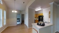This single-story space showcases an open and functional layout that seamlessly connects the kitchen and living areas. The kitchen is outfitted with warm honey-toned cabinetry, a black refrigerator, and light countertops, with fluorescent overhead lighting ensuring a bright workspace. A white peninsula extends into the common area, offering additional counter space and a natural division between the kitchen and the rest of the home.

Hardwood-style flooring runs throughout, lending warmth and continuity to the space. The vaulted ceiling in the living area creates an airy, elevated feel, further enhanced by a classic chandelier that adds a touch of elegance. Crown molding accents the ceiling transitions, contributing to the overall refined aesthetic.

A central hallway provides access to additional rooms, including what appears to be a bedroom visible through an open doorway, suggesting a well-organized floor plan. A closet with bi-fold doors offers convenient storage along the hall. The neutral gray wall tones throughout create a cohesive, understated backdrop that complements the natural wood flooring and cabinetry.