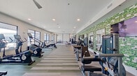 This expansive fitness center is designed to support a wide range of workout styles and training goals. The main exercise floor is finished with a distinctive striped multi-tone carpet, providing a clean, contemporary aesthetic throughout. Large windows line the perimeter walls, flooding the space with natural light and offering views of an adjacent outdoor area, including what appears to be a pool deck with a pergola structure.

The cardio zone is well-equipped with treadmills, elliptical trainers, stationary bikes, rowing machines, and stair climbers, all arranged to maximize space and comfort. A dedicated spinning area features a row of cycling bikes positioned near the windows. Strength training is supported by a selection of cable machines, weight benches, and free weights neatly organized along a striking green textured brick accent wall, complemented by large mirrors that enhance the sense of depth.

A separate functional training room features wood-look flooring, a punching bag, multi-functional training rack, medicine balls, and resistance equipment against a bold deep-red accent wall. Ceiling fans and recessed lighting are installed throughout, ensuring a comfortable environment. The overall layout and variety of equipment make this a thorough, well-appointed fitness facility.