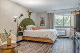 Located at 517 Grand Canyon Dr in Madison, Wisconsin, this well-appointed one-bedroom, two-bathroom suite offers a thoughtfully designed, efficient layout across a single floor. Light wood-tone flooring runs throughout the main living area, creating a warm and cohesive aesthetic. The bedroom area is the centerpiece of the space, showcasing a statement platform bed with an arched green velvet headboard, complemented by eclectic décor including a sculptural wall mount and modern multi-arm floor lamp. A cozy accent chair adds a comfortable lounge area alongside the bed. Natural light filters in through a large window with floor-length drapes, offering views of the surrounding greenery. A built-in kitchenette lines one wall, featuring dark cabinetry, a stainless steel refrigerator, microwave, and a sink — providing practical in-room convenience. A wall-mounted flat-screen TV and a light wood dresser complete the living area. The entry corridor leads to two separate bathrooms, each finished with neutral tile flooring. One bathroom features a shower-tub combo with a curtain, toilet, and towel bar, while a shared vanity area with a long countertop and mirror serves both spaces efficiently. The overall ambiance is modern yet inviting, blending functional design with stylish, curated furnishings.