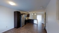 This two-floor space offers a thoughtfully designed layout that balances functionality with modern aesthetics. The open-concept main level showcases a sleek kitchen equipped with stainless steel appliances, including a full-sized refrigerator, microwave, and dishwasher. Rich, dark cabinetry provides generous storage and creates a striking contrast against the neutral-toned walls and warm hardwood flooring that flows seamlessly throughout the space. A kitchen peninsula with pendant lighting serves as a natural dividing point between the kitchen and the adjoining living area, enhancing both the visual appeal and practicality of the layout. Track lighting above the kitchen adds focused illumination, while a large window fitted with vertical blinds invites natural light into the living area, creating a bright and welcoming ambiance. A glass-paneled door leads to what appears to be an outdoor area, further enhancing the sense of openness and connectivity to the exterior. The clean lines, contemporary finishes, and cohesive design elements across both floors contribute to a polished, move-in-ready environment suited for comfortable everyday living.