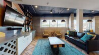 Located at 703 Washington Ave in Towson, Maryland, this single-floor space delivers a compelling blend of industrial and contemporary design. The layout opens into a spacious, light-filled lounge anchored by striking herringbone-patterned hardwood flooring that transitions seamlessly into large-format slate tile throughout the entryway corridor. A bold navy-painted exposed ceiling with industrial pipe detailing and track lighting sets a dramatic tone overhead, while oversized pendant dome fixtures provide warm, focused illumination throughout the seating areas. The lounge area is thoughtfully furnished with a rich leather sofa accented by teal and mustard throw pillows, a pair of upholstered arm chairs, and a weathered wood coffee table atop a vibrant teal area rug. A built-in media console with white cabinetry, decorative cinder block accents, and a mounted flat-screen TV is set against a bold geometric mural, flanked by an industrial pipe-style shelving unit. Natural light pours in through a row of wide windows framing views of the surrounding neighborhood. A secondary lounge zone features vivid yellow and blue walls adorned with colorful artwork, offering a lively contrast to the darker main lounge. The entry corridor includes a built-in bench with cushioned seating, leading toward a bright dining area beyond.