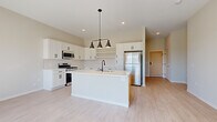 This well-appointed single-floor unit showcases a clean, contemporary design with light wood-look flooring and neutral tones carried consistently throughout the space. The heart of the unit is the open-concept kitchen, which features crisp white shaker cabinetry with matte black hardware, a white quartz countertop, and a subway tile backsplash. A center island with a warm wood base provides additional prep space and includes a built-in sink with a matte black faucet, complemented by a stylish three-light pendant fixture overhead. Stainless steel appliances — including a gas range, over-the-range microwave, and refrigerator — complete the kitchen. Open shelving adds a functional, modern touch alongside the upper cabinetry. The living area receives generous natural light through a full sliding glass door that opens to a private balcony, offering outdoor views of a landscaped green space. The bathroom features a dual-sink vanity with white cabinetry, matte black fixtures, quartz countertops, and a large mirror. A dedicated in-unit laundry area houses a full-size side-by-side washer and dryer with overhead wire shelving. Additional rooms are bright and well-proportioned, each featuring the same light flooring and neutral wall finishes, offering a versatile and move-in-ready layout.