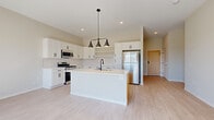 This well-appointed single-floor unit showcases a clean, contemporary design with light wood-look flooring and neutral tones carried consistently throughout the space. The heart of the unit is the open-concept kitchen, which features crisp white shaker cabinetry with matte black hardware, a white quartz countertop, and a subway tile backsplash. A center island with a warm wood base provides additional prep space and includes a built-in sink with a matte black faucet, complemented by a stylish three-light pendant fixture overhead. Stainless steel appliances — including a gas range, over-the-range microwave, and refrigerator — complete the kitchen. Open shelving adds a functional, modern touch alongside the upper cabinetry. The living area receives generous natural light through a full sliding glass door that opens to a private balcony, offering outdoor views of a landscaped green space. The bathroom features a dual-sink vanity with white cabinetry, matte black fixtures, quartz countertops, and a large mirror. A dedicated in-unit laundry area houses a full-size side-by-side washer and dryer with overhead wire shelving. Additional rooms are bright and well-proportioned, each featuring the same light flooring and neutral wall finishes, offering a versatile and move-in-ready layout.