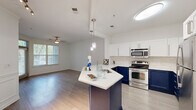 This thoughtfully designed single-floor residence offers a cohesive open-concept layout that connects the kitchen, dining, and living areas into one functional and inviting space. The kitchen is a standout feature, showcasing striking two-tone cabinetry with bold navy lower cabinets paired with crisp white upper cabinets, complemented by a classic subway tile backsplash and white quartz countertops. A central island with pendant lighting serves as both a prep space and casual dining area, while stainless steel appliances complete the modern aesthetic. Light hardwood-style flooring flows throughout the main living areas, brightened by natural light streaming through slatted window blinds and accented by crown molding details. The living room is spacious and open, featuring a ceiling fan and clean white walls that create a neutral, versatile canvas. The bedroom offers plush carpet flooring, crown molding, a ceiling fan, and a sun-lit window, providing a comfortable and private retreat. The bathroom is finished with a wide vanity, white cabinetry with brushed nickel hardware, a large mirror with overhead lighting, and light tile flooring. A hallway and closet round out the practical layout, ensuring efficient use of every square foot in this well-planned residence.