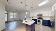 This thoughtfully designed single-floor residence offers a cohesive open-concept layout that connects the kitchen, dining, and living areas into one functional and inviting space. The kitchen is a standout feature, showcasing striking two-tone cabinetry with bold navy lower cabinets paired with crisp white upper cabinets, complemented by a classic subway tile backsplash and white quartz countertops. A central island with pendant lighting serves as both a prep space and casual dining area, while stainless steel appliances complete the modern aesthetic. Light hardwood-style flooring flows throughout the main living areas, brightened by natural light streaming through slatted window blinds and accented by crown molding details. The living room is spacious and open, featuring a ceiling fan and clean white walls that create a neutral, versatile canvas. The bedroom offers plush carpet flooring, crown molding, a ceiling fan, and a sun-lit window, providing a comfortable and private retreat. The bathroom is finished with a wide vanity, white cabinetry with brushed nickel hardware, a large mirror with overhead lighting, and light tile flooring. A hallway and closet round out the practical layout, ensuring efficient use of every square foot in this well-planned residence.
