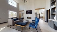 Located at 34 Bank St in Netcong, New Jersey, this single-floor community clubhouse delivers a sophisticated, contemporary design with a variety of thoughtfully appointed spaces. The centerpiece is a grand lounge area distinguished by dramatically soaring ceilings, oversized windows that flood the room with natural light, and a sleek linear fireplace set against a glossy white accent wall. A wall-mounted flat-screen TV and a striking geometric pendant chandelier complete the room, while plush seating—including a navy velvet ottoman, cobalt blue accent chairs, and a grey sectional sofa—adds a refined sense of comfort atop bold striped area rugs and light wood-look tile flooring. Adjacent to the lounge is a well-designed co-working station featuring built-in shelving, a large-format artistic mural, task seating, and ample desk space. The game room offers a billiards table, a mounted TV, decorative slatted wood partitions, and casual bar-height seating. A spacious fitness or flex room provides light wood floors and multiple windows overlooking the surrounding community. The kitchen area stands out with navy cabinetry, gold-accented open shelving, a central island with bar seating, and geometric wire pendant lights, all beneath soaring ceilings with abundant natural light.