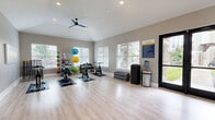 Located at 13872 Del Corso Way in Broomfield, Colorado, this dedicated exercise room is a thoughtfully designed fitness studio built for group classes and active training. The space features vaulted ceilings that lend an airy, open atmosphere, complemented by light wood-look flooring that extends throughout the room. Multiple large windows fitted with horizontal blinds line the walls, flooding the interior with abundant natural light and offering views of the surrounding outdoor greenery. A sleek black ceiling fan adds to the comfortable climate of the room.

The studio is outfitted with a row of stationary cycling bikes positioned on protective floor mats, making it well-suited for spin classes. A freestanding equipment rack holds an assortment of colorful stability balls and resistance tools, while a stack of exercise mats provides options for floor-based workouts. A full-length wall mirror on one end enhances the sense of space and supports form-checking during exercise. A mounted flat-screen display is also present, ideal for guided instruction.

Glass-paneled double doors provide easy access to adjacent areas and maintain a bright, connected feel. Studio rules are posted near the entrance, reinforcing the space's organized, community-oriented design. Overall, the studio offers a clean, modern, and energizing environment well-suited for a variety of fitness pursuits.