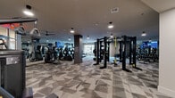 Located at 901 St Albans Dr in Raleigh, NC, this well-appointed fitness center offers a generously sized, single-floor exercise space designed to support a comprehensive range of workout routines. The room is finished with a striking geometric-patterned carpet in neutral gray tones, lending a modern, polished aesthetic throughout.

The cardio zone features a row of treadmills and elliptical machines, each equipped with individual digital screens, alongside stationary and spin bikes. Ceiling fans and recessed lighting ensure a comfortable, well-lit environment. Large windows along one wall invite natural light and provide an open, airy feel.

The strength training area is equally well-equipped, housing a multi-station power rack system with TRX suspension trainers, cable machines, and a full dumbbell rack. A dedicated functional training alcove includes medicine balls, stability balls, kettlebells, and a heavy punching bag, catering to dynamic and high-intensity workouts. Floor-to-ceiling mirrors line multiple walls, amplifying the sense of space and supporting proper form.

A separate nook area near the entrance provides additional storage and beverage refrigeration. Motivational artwork accents the walls, contributing to an energetic atmosphere. The space is thoughtfully laid out to allow for smooth traffic flow between zones, making it a fully equipped destination for fitness at every level.