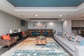 This single-floor amenity space is thoughtfully designed with a contemporary aesthetic that blends bold color accents with sophisticated, neutral finishes throughout. The open-concept kitchen is a standout feature, boasting floor-to-ceiling soft-gray cabinetry, a richly tiled backsplash, stainless steel appliances including a double wall oven and French door refrigerator, and a striking teal island topped with speckled granite countertops. Recessed lighting and a geometric gold pendant fixture illuminate the space with warmth. Adjacent to the kitchen, a dedicated wet bar area features a draft beer tap, wine storage, and a built-in beverage cooler framed by a dramatic copper-toned mosaic tile accent wall, with a pendant light adding a cozy glow. The expansive lounge area serves as the social heart of the space, anchored by a vivid, multi-toned area rug and a geometric wood coffee table. Seating includes plush sofas, leather accent chairs, and a built-in banquette dining nook with mid-century modern chairs. A tray ceiling painted in deep teal and burnt orange adds a bold architectural focal point overhead. A wall-mounted flat-screen TV, textured wallcovering, and curated artwork contribute to the polished, community-oriented ambiance. Floor-to-ceiling glass doors open directly to an outdoor patio, flooding the lounge with natural light and seamlessly extending the living experience outdoors.