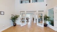 The Springwater Apartments clubhouse at 1205 Colgin Dr in Virginia Beach, Virginia, is a thoughtfully designed community amenity space that combines elegance with functionality. The entry makes an immediate impression with soaring double-height ceilings, floor-to-ceiling glass French doors, and warm tile flooring that flows throughout the entire facility. Flanking the entrance, tall potted palms add a welcoming, resort-like atmosphere.

Beyond the grand entry vestibule, the space opens into several distinct yet interconnected areas. A bright, airy dining nook features classic wainscoting, large windows overlooking the outdoor courtyard, and a round communal table with wishbone chairs. Adjacent to this is a striking kitchen and bar area outfitted with slate-blue cabinetry, quartz countertops, a geometric tile backsplash, and a wall-mounted television — ideal for community gatherings and events.

The lounge area offers comfortable, tufted leather seating, a large flat-screen TV, and views into neighboring spaces through expansive glass windows, creating a connected yet intimate feel. A dedicated conference room provides a professional setting with a wood-top table, ergonomic seating, and natural light from a large window with garden views. Throughout, the design palette of crisp whites, soft blues, and natural wood tones creates a cohesive, coastal-inspired ambiance.