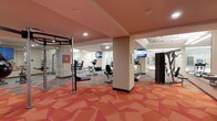 This fitness center offers a thoughtfully designed workout space spread across a single level. The room is anchored by a striking geometric-patterned carpet in bold shades of coral, red, and gray, lending the space a modern and energetic aesthetic that complements its function. The ceiling features a warm salmon-toned finish with recessed lighting, while a decorative ornate ceiling detail is visible in the far section of the room, adding an unexpected architectural touch.

The space is divided into distinct workout zones by a central structural column. The cardio area hosts multiple treadmills and upright exercise bikes, each equipped with individual screens for entertainment during workouts. Wall-mounted flat-screen TVs are positioned throughout, ensuring visibility from various equipment locations. A dedicated strength training section includes a multi-functional rack with accessories and additional resistance equipment. A glass-enclosed room along one wall provides a private workout or stretching area, enhanced by colorful wall art. The overall ambiance is bright, clean, and motivating, making it a functional and visually engaging facility.