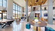 Located at 2515 S Verde Dr in Gilbert, AZ, this impressive two-story amenity space offers a generously scaled environment designed for both comfort and social engagement. The ground floor welcomes visitors with a soaring double-height lobby, featuring floor-to-ceiling curtain-wall windows that flood the space with natural Arizona sunlight and provide sweeping outdoor views. A slatted wood ceiling accent, polished concrete floors, dramatic pendant lighting, and a striking central round table with teal ottomans establish a refined, contemporary tone. A whitewashed brick accent wall frames a large-screen TV and open shelving, anchoring a relaxed lounge area furnished with leather sofas and accent chairs. A shuffle board table and bar-height seating along the brick wall add a social, recreational dimension. A sculptural open-tread staircase with a warm wood and black metal railing connects the floors with architectural elegance. The upper floor expands into wide, light-filled corridors with carpeted flooring, fluted gray columns, and mezzanine views overlooking the lobby below. Multiple seating vignettes offer flexible gathering spaces, including a lounge with a whitewashed brick media wall, a window-side bench with pendant lighting and a slatted wood divider screen, and intimate seating areas with large windows framing community views.