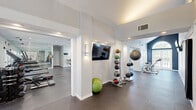 This fitness center offers a spacious, thoughtfully designed workout facility spread across a single floor. The space is divided into two distinct functional zones, each catering to different aspects of physical training. The interior features a clean, modern aesthetic with crisp white walls, recessed lighting, and durable rubber flooring throughout, creating a professional and motivating environment.

The cardio zone is equipped with treadmills, an elliptical, and a rowing machine, all positioned near large windows that invite ample natural light and offer views of the surrounding greenery. Flat-screen televisions are mounted overhead for entertainment during workouts. The strength training area houses a dedicated Training Zone multi-functional cable machine, a variety of selectorized weight machines, a medicine ball rack, stability balls, and a stationary cycle.

Floor-to-ceiling mirrors are strategically placed throughout to enhance the sense of space and allow users to monitor their form. The facility is accessed through elegant glass-paneled double doors framed by soaring ceilings, which lend an airy, open feel to the entrance. Multiple entry and exit points provide convenient access. The overall ambiance is bright, clean, and well-organized, making this fitness center a fully equipped destination for a comprehensive workout experience.