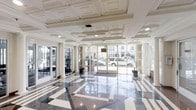 This elegant single-floor lobby makes a strong architectural impression through its carefully considered design elements. The floor is finished in polished terrazzo, featuring a bold geometric pattern of dark and light tones that reflects the ambient light and adds visual depth to the generous open layout. Overhead, an ornate coffered ceiling with layered molding detailing lends a classical, formal character to the space.

Floor-to-ceiling glass walls and wide glass entry doors line the front of the lobby, allowing abundant natural light to pour in and providing clear sightlines to the exterior streetscape. White structural columns punctuate the perimeter, reinforcing the neoclassical aesthetic while contributing to the sense of openness and height.

Potted plants positioned near the entry doors introduce a subtle organic element, softening the otherwise crisp, formal palette of whites and grays. To one side, fitness equipment is partially visible, suggesting an ancillary amenity area integrated into the layout. The overall ambiance is clean, refined, and professional, combining classic architectural detailing with bright, well-lit interiors to create a welcoming and polished environment.