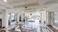 This elegant single-floor lobby makes a strong architectural impression through its carefully considered design elements. The floor is finished in polished terrazzo, featuring a bold geometric pattern of dark and light tones that reflects the ambient light and adds visual depth to the generous open layout. Overhead, an ornate coffered ceiling with layered molding detailing lends a classical, formal character to the space.

Floor-to-ceiling glass walls and wide glass entry doors line the front of the lobby, allowing abundant natural light to pour in and providing clear sightlines to the exterior streetscape. White structural columns punctuate the perimeter, reinforcing the neoclassical aesthetic while contributing to the sense of openness and height.

Potted plants positioned near the entry doors introduce a subtle organic element, softening the otherwise crisp, formal palette of whites and grays. To one side, fitness equipment is partially visible, suggesting an ancillary amenity area integrated into the layout. The overall ambiance is clean, refined, and professional, combining classic architectural detailing with bright, well-lit interiors to create a welcoming and polished environment.