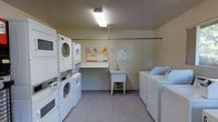 This communal laundry facility is a dedicated, single-room space designed to serve residents with a practical and functional layout. The room is outfitted with multiple stacked washer-dryer units along one wall, maximizing the use of vertical space, while additional standalone top-load washers and dryers are positioned along the opposite side, providing ample capacity for simultaneous use. A freestanding utility sink at the center of the room offers a convenient station for hand-washing or pre-treating garments. A built-in folding counter along the back wall provides a dedicated surface for sorting and folding laundry. A community bulletin board is mounted on the wall, displaying notices and guidelines for residents, including rules regarding adult supervision and pet restrictions. The room is illuminated by overhead fluorescent lighting, ensuring a bright and well-lit environment. Light-toned flooring and neutral wall colors contribute to a clean, utilitarian aesthetic. The space is thoughtfully organized to accommodate multiple users at once, making it a highly functional shared amenity.