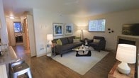 This single-floor space features a thoughtfully designed living area that balances comfort and contemporary style. The living room is anchored by warm hardwood flooring that extends throughout, providing a cohesive and inviting foundation. A large, plush area rug defines the seating area, which centers around a dark gray sofa accented with green and patterned throw pillows, complemented by a matching armchair. A sleek glass-top coffee table adds a modern touch to the arrangement.

The walls are painted in a clean, neutral tone, creating a bright and airy ambiance. Two framed abstract artworks with blue and green hues add a splash of color and personality to the space. Ambient lighting is provided by a pair of floor and table lamps, casting a warm glow throughout the room. A window overlooking lush greenery allows natural light to filter in, enhancing the open feel of the space.

Additional details include a small side table with decorative accents and a wall-mounted heater vent, contributing to the room's functional yet stylish character. The overall layout is efficient and well-organized, making excellent use of the available space.