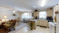 This single-floor residence offers a warm and inviting open-plan layout that seamlessly connects the kitchen and dining areas, creating a cohesive living environment ideal for everyday comfort and casual entertaining. The kitchen is well-appointed with cream-toned cabinetry, a central island with bar-height seating, and a double sink positioned beneath a window that draws in natural light. A skylight overhead further brightens the space, complementing the light tile flooring that extends throughout the kitchen area. Ample counter space and storage make the kitchen both functional and practical. The adjacent dining area features a wooden table with matching chairs set atop plush carpeting, adding a cozy contrast to the tiled kitchen zone. A ceiling fan with a light fixture keeps the space comfortable and airy. Multiple windows dressed with sheer curtains and decorative drapes allow generous amounts of natural light to fill the room while maintaining a soft, homey atmosphere. Thoughtful decorative touches — including a wreath, potted plants, and framed wall art — lend the space a personal, lived-in character. The overall ambiance is light, clean, and welcoming, reflecting a well-maintained and comfortably arranged home.