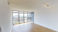 This single-floor high-rise unit offers a sleek, contemporary living environment defined by its most striking feature: expansive floor-to-ceiling windows that wrap around the corner of the main living area. These oversized windows flood the space with abundant natural light throughout the day and frame panoramic views of the surrounding urban skyline, neighboring residential buildings, and a lush tree canopy stretching into the distance — a rare combination of city energy and natural scenery.

The interior is finished with warm-toned hardwood flooring that contrasts beautifully against the crisp white walls and ceilings, creating a clean and inviting aesthetic. The open layout of the living area maximizes the sense of space and airiness, with the corner window configuration enhancing the feeling of openness from multiple vantage points.

The neutral color palette throughout provides a versatile backdrop, making the space adaptable to a wide range of interior design styles. The elevated position of the unit enhances privacy while delivering unobstructed sightlines across the cityscape. Overall, the space combines modern architectural design with practical, light-filled interiors, making it a well-appointed urban residence with a strong connection to its surrounding environment.