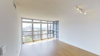 This single-floor high-rise unit offers a sleek, contemporary living environment defined by its most striking feature: expansive floor-to-ceiling windows that wrap around the corner of the main living area. These oversized windows flood the space with abundant natural light throughout the day and frame panoramic views of the surrounding urban skyline, neighboring residential buildings, and a lush tree canopy stretching into the distance — a rare combination of city energy and natural scenery.

The interior is finished with warm-toned hardwood flooring that contrasts beautifully against the crisp white walls and ceilings, creating a clean and inviting aesthetic. The open layout of the living area maximizes the sense of space and airiness, with the corner window configuration enhancing the feeling of openness from multiple vantage points.

The neutral color palette throughout provides a versatile backdrop, making the space adaptable to a wide range of interior design styles. The elevated position of the unit enhances privacy while delivering unobstructed sightlines across the cityscape. Overall, the space combines modern architectural design with practical, light-filled interiors, making it a well-appointed urban residence with a strong connection to its surrounding environment.