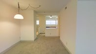 This single-floor residence offers a straightforward and functional layout designed for comfortable everyday living. The main living area is finished with neutral wall-to-wall carpeting that lends a soft, understated feel to the space, while light-toned walls and standard ceiling height create a clean, open atmosphere. A decorative chain-hung light fixture adds a modest focal point to the living area.

The kitchen is situated toward the rear of the open-plan space and features a half-wall pass-through that connects it visually to the living area, enhancing the sense of openness. The kitchen is equipped with a sink positioned beneath a window that allows natural light to filter in, brightening both the kitchen and the adjacent areas. Warm-toned cabinetry provides practical storage space, and a flush-mount ceiling light ensures adequate illumination.

A hallway visible beyond the kitchen suggests additional rooms beyond the main living space, providing privacy and separation for sleeping or utility areas. The overall design of the residence prioritizes functionality and simplicity, with consistent neutral finishes throughout. Ventilation is supported by ceiling vents, and the layout is arranged to maximize the usability of the available floor space across its single level.