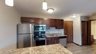 This single-floor space centers around a thoughtfully designed kitchen that combines functionality with a warm, inviting aesthetic. The kitchen is outfitted with a full suite of stainless steel appliances, including a refrigerator, electric range with oven, over-the-range microwave, and dishwasher. Rich, dark-toned wood cabinetry lines the walls, offering generous upper and lower storage with a clean, shaker-style design. The countertops feature granite with warm gold and brown tones, adding a touch of elegance to the space. A kitchen island with matching granite provides additional workspace and casual seating potential. Pendant and flush-mount ceiling fixtures illuminate the kitchen with layered lighting. The flooring transitions from light hardwood-style planks in the kitchen area to the adjacent spaces, contributing to a cohesive look throughout. A wood-paneled door visible in the background adds a natural element, complementing the cabinetry tones. Overall, the space presents a clean, modern interior with warm finishes that create a comfortable and practical living environment.