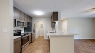 This single-floor, 1-bedroom, 1-bathroom unit offers a thoughtfully designed layout that maximizes comfort and functionality. The kitchen is a standout feature, showcasing dark wood cabinetry, quartz countertops, a subway tile backsplash, and a full suite of stainless steel appliances including a gas range, microwave, and French door refrigerator. A peninsula with a single-basin sink provides additional prep space and opens to the living area, creating an airy, connected feel. Warm hardwood-style flooring flows throughout the main living spaces, adding a welcoming ambiance. A pendant light fixture and recessed lighting keep the kitchen and living area bright and inviting. The spacious bedroom features plush carpeting, a ceiling fan, and a window that brings in natural light and outdoor views, complemented by a dedicated closet for storage. The bathroom is finished with light wood cabinetry, a quartz-top vanity, a lighted mirror, and a tub-shower combination. A dedicated in-unit laundry area with a full-size top-load washer and dryer, topped with additional cabinetry, adds everyday convenience. Neutral tones and clean finishes throughout give the space a modern, move-in-ready appeal.