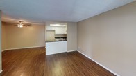 This single-floor residence offers a well-organized layout with distinct living spaces, each finished with practical materials and neutral warm tones throughout. The living area is generously sized, featuring rich dark hardwood-style flooring, a textured ceiling, and a window fitted with blinds that invites natural light and outdoor views. A pass-through opening connects the living space directly to the kitchen, enhancing the open feel between the two areas. The kitchen is efficiently designed with a U-shaped layout, featuring white cabinetry, dark speckled countertops, a black electric range with overhead microwave, a double stainless steel sink, and hardwood-style flooring. A recessed tray ceiling with a flush-mount light fixture adds a refined touch overhead. The bedroom is carpeted with plush flooring, painted in a matching warm beige tone, and includes a wide sliding-door closet offering ample storage with a full hanging rod. A window with blinds provides natural light and ventilation. The bathroom features a full-size soaking tub with an overhead shower and chrome fixtures, set within a clean white surround. Throughout the space, textured ceilings and consistent neutral wall tones create a cohesive, move-in ready environment.