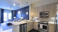 This single-floor space showcases a thoughtfully designed open-concept layout that seamlessly connects the kitchen and living area. The kitchen is a standout feature, equipped with stainless steel appliances including a range, built-in microwave, and dishwasher. Light wood cabinetry with sleek hardware provides ample storage, while the white quartz countertops offer generous prep space. A textured tile backsplash adds visual interest behind the range, complementing the overall modern aesthetic. An L-shaped peninsula with an undermount sink doubles as a breakfast bar, creating a natural transition into the living space. Pendant lighting hangs above the peninsula, while a multi-directional track lighting system illuminates the kitchen and adjacent areas. The bold navy blue accent wall in the living area creates a striking focal point, adding depth and a contemporary edge to the space. The living area is open and inviting, with décor that echoes the cool, modern color palette. Together, the spaces feel cohesive and well-curated, with a design language that balances function and style throughout.