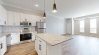 This thoughtfully designed one-bedroom, one-bathroom residence offers a clean, modern aesthetic across its single floor. The open-concept kitchen and living area serves as the heart of the home, anchored by a granite-topped center island illuminated by sleek pendant lighting. The kitchen is outfitted with crisp white shaker cabinets accented by matte black hardware, a white subway tile backsplash, and a full suite of stainless steel appliances including a refrigerator, range, microwave, and dishwasher. Warm-toned hardwood-style flooring flows throughout the main living areas, complementing the soft gray walls and crown molding that lend an elevated finish to the space. The bedroom offers a comfortable retreat with plush carpeting, natural light from a window with blinds, and a double-door closet providing ample storage. A conveniently located in-unit washer and dryer adds everyday practicality. The bathroom is finished with a granite-topped white vanity, a framed mirror, modern light fixture, and a full soaking tub with shower. Together, the neutral palette, quality finishes, and functional layout create a cohesive and inviting living environment.