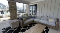 This covered outdoor lounge space offers a seamless blend of comfort and contemporary design. The area is anchored by a large wicker sectional sofa dressed in plush light grey cushions and accented with colorful throw pillows, arranged around a slatted wood coffee table and a bold black-and-white patterned area rug. Board-and-batten style wall paneling in a soft grey tone lines one side of the space, adding a clean, modern aesthetic. A striking natural stone column serves as a focal architectural element, complementing the exterior materials and adding texture and warmth. Floor-to-ceiling screened openings span the perimeter, flooding the lounge with abundant natural light while maintaining a sheltered, comfortable environment in all seasons. A set of sleek black French doors connects the lounge to the interior, providing an elegant transition between indoor and outdoor living. Views through the screens reveal a well-maintained surrounding community with residential buildings and a pool area, enhancing the sense of openness. The overall ambiance is refined yet relaxed, making this space well-suited for both casual lounging and social gatherings.