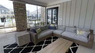 This covered outdoor lounge space offers a seamless blend of comfort and contemporary design. The area is anchored by a large wicker sectional sofa dressed in plush light grey cushions and accented with colorful throw pillows, arranged around a slatted wood coffee table and a bold black-and-white patterned area rug. Board-and-batten style wall paneling in a soft grey tone lines one side of the space, adding a clean, modern aesthetic. A striking natural stone column serves as a focal architectural element, complementing the exterior materials and adding texture and warmth. Floor-to-ceiling screened openings span the perimeter, flooding the lounge with abundant natural light while maintaining a sheltered, comfortable environment in all seasons. A set of sleek black French doors connects the lounge to the interior, providing an elegant transition between indoor and outdoor living. Views through the screens reveal a well-maintained surrounding community with residential buildings and a pool area, enhancing the sense of openness. The overall ambiance is refined yet relaxed, making this space well-suited for both casual lounging and social gatherings.
