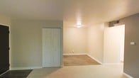 This single-floor space presents a clean, functional layout with a neutral color palette throughout. The interior features warm beige walls and white trim, creating a bright and cohesive aesthetic. A notable design element is the transition between two distinct flooring types — plush carpeting in one area and smooth hardwood-style flooring in an adjacent section, offering both comfort and practicality depending on the use of each zone. A six-panel closet door provides convenient built-in storage, making efficient use of the available space. Overhead, a flush-mount ceiling light fixture provides consistent ambient illumination throughout the room. White baseboards run along the perimeter, adding a crisp, finished look to the walls. An electrical outlet is visible along the baseboard, indicating the space is ready for immediate furnishing and use. The overall ambiance is understated and move-in ready, with a simple, uncluttered design that provides a versatile backdrop suitable for a variety of uses and personal styles.