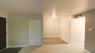 This single-floor space presents a clean, functional layout with a neutral color palette throughout. The interior features warm beige walls and white trim, creating a bright and cohesive aesthetic. A notable design element is the transition between two distinct flooring types — plush carpeting in one area and smooth hardwood-style flooring in an adjacent section, offering both comfort and practicality depending on the use of each zone. A six-panel closet door provides convenient built-in storage, making efficient use of the available space. Overhead, a flush-mount ceiling light fixture provides consistent ambient illumination throughout the room. White baseboards run along the perimeter, adding a crisp, finished look to the walls. An electrical outlet is visible along the baseboard, indicating the space is ready for immediate furnishing and use. The overall ambiance is understated and move-in ready, with a simple, uncluttered design that provides a versatile backdrop suitable for a variety of uses and personal styles.