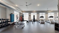Located at 600 NW Cherrycreek Ln in Ankeny, Iowa, this fitness center offers a generously sized, single-floor exercise space designed to accommodate a comprehensive workout experience. The facility is divided into two distinct functional zones seamlessly connected within an open layout.

The cardio area lines one side of the room and features a row of treadmills, elliptical trainers, stair climbers, and a rowing machine. Multiple flat-screen TVs are mounted on the textured concrete block wall, providing entertainment during workouts. Ceiling fans throughout the space ensure consistent air circulation, complementing the recessed lighting overhead. Roller shade-equipped windows along the perimeter allow ample natural light while maintaining privacy.

The strength training zone offers a full selection of free weights, including multiple dumbbell racks, adjustable benches, medicine balls, a cable machine, and a dedicated stretching and floor exercise area. Large wall mirrors in the free weights section enhance spatial awareness during training. Durable rubber flooring covers the entire space, providing both safety and comfort.

Additional amenities include a mini-refrigerator, storage cubbies, and a waste receptacle station along the wall. Exposed ductwork along the ceiling adds an industrial aesthetic touch. The overall design is practical, clean, and well-organized, making this facility a fully functional fitness destination.